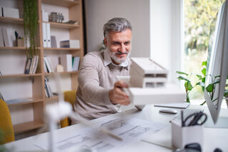AdobeStock_320587708 Older architect, with house model, desk, office, working